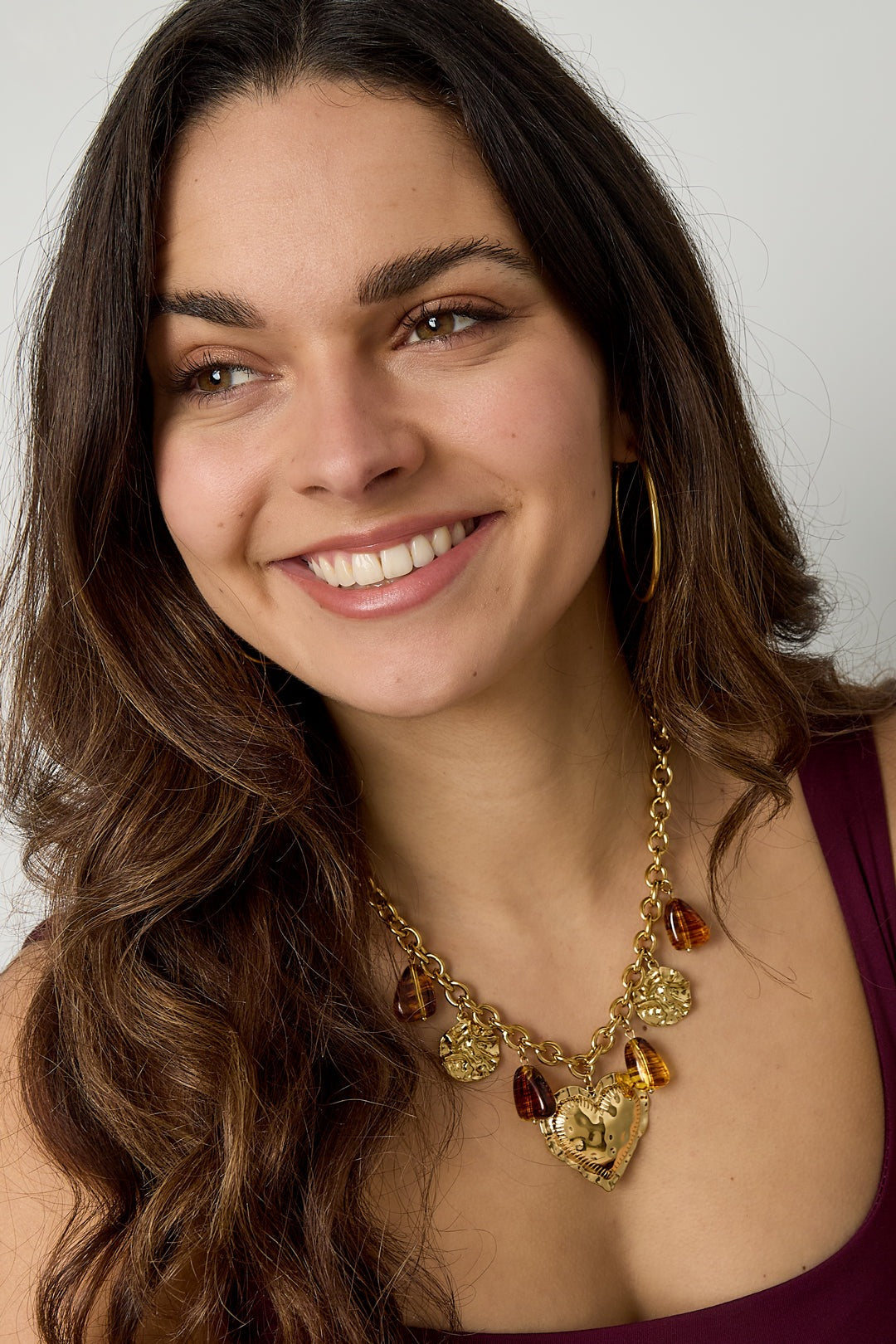 Smiling woman wearing chunky gold-tone necklace with hammered heart charm and amber resin accents paired with hoop earrings and sleeveless top