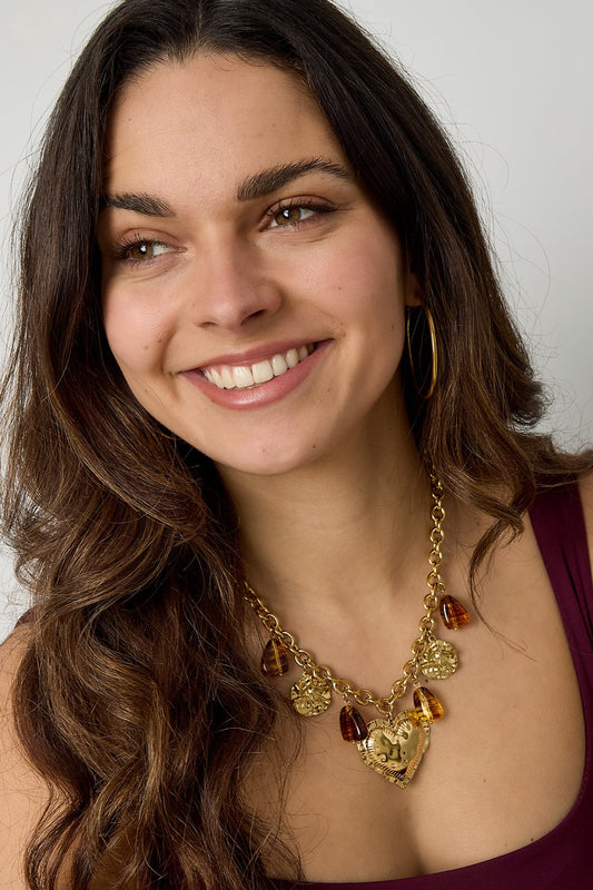 Smiling woman wearing chunky gold-tone necklace with hammered heart charm and amber resin accents paired with hoop earrings and sleeveless top