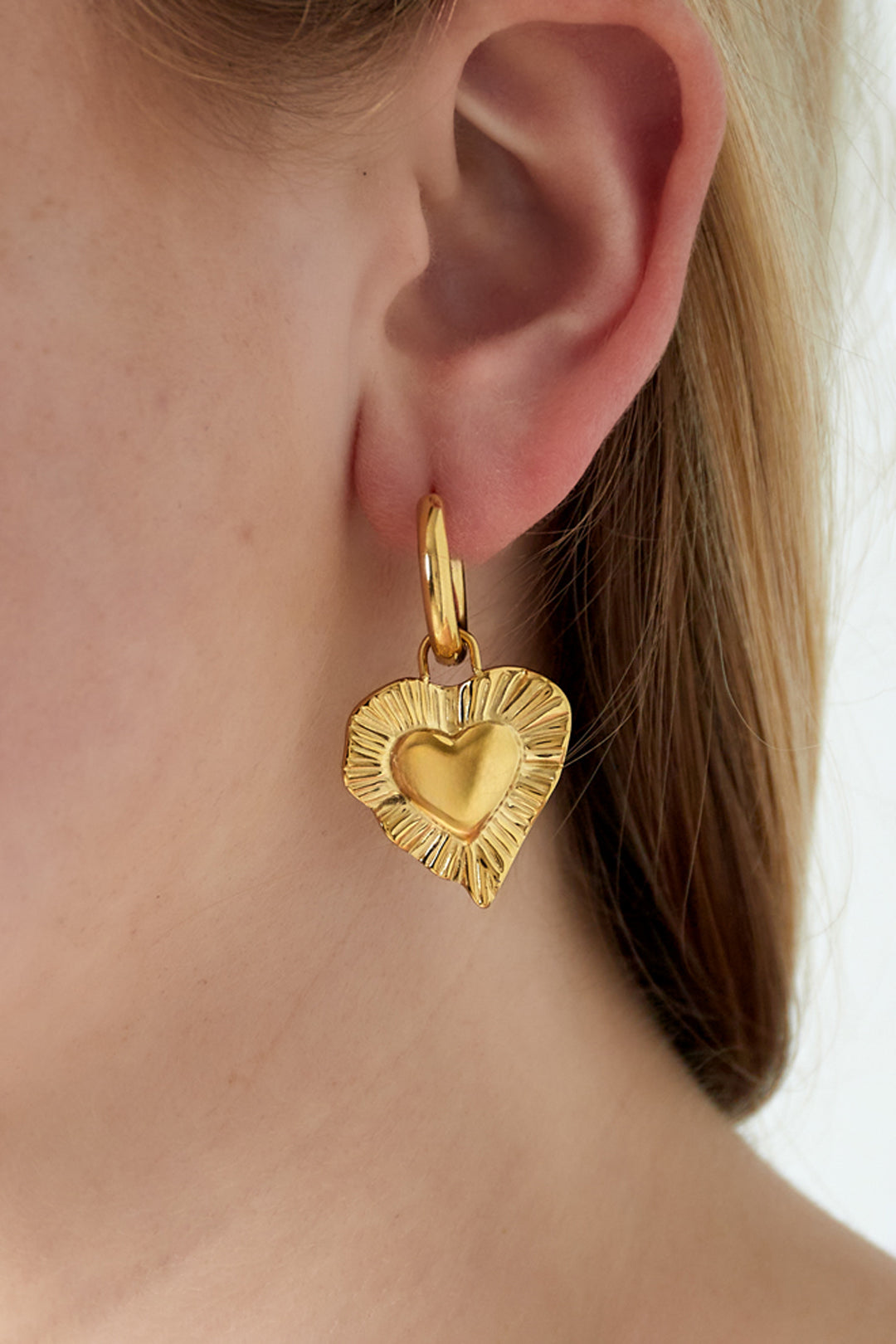 Close-up of a woman's ear wearing a gold radiant heart charm drop earring with smooth hoop fastening and textured heart pendant.