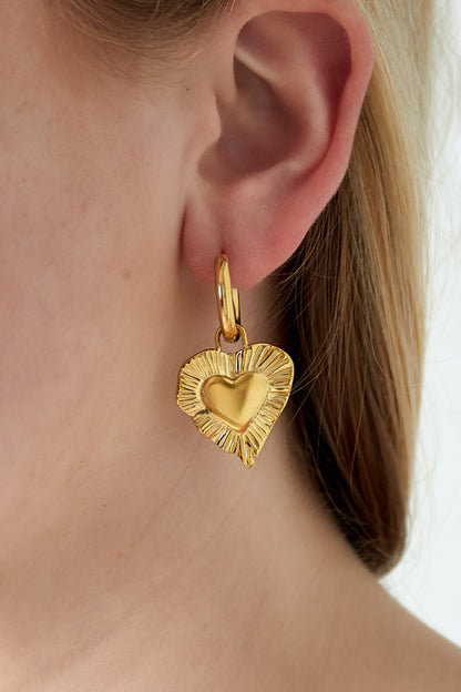 Close-up of a woman's ear wearing a gold radiant heart charm drop earring with smooth hoop fastening and textured heart pendant.