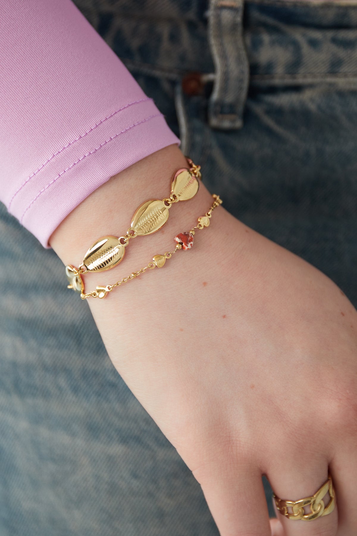 Close-up of a wrist adorned with the gold shell charm bracelet paired with a delicate gold charm chain bracelet, worn with casual denim and pink sleeve
