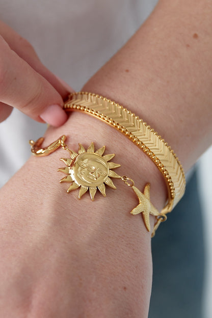 Close-up of gold celestial charm bracelet worn on wrist alongside textured gold bangles, showing details of sun and star charms