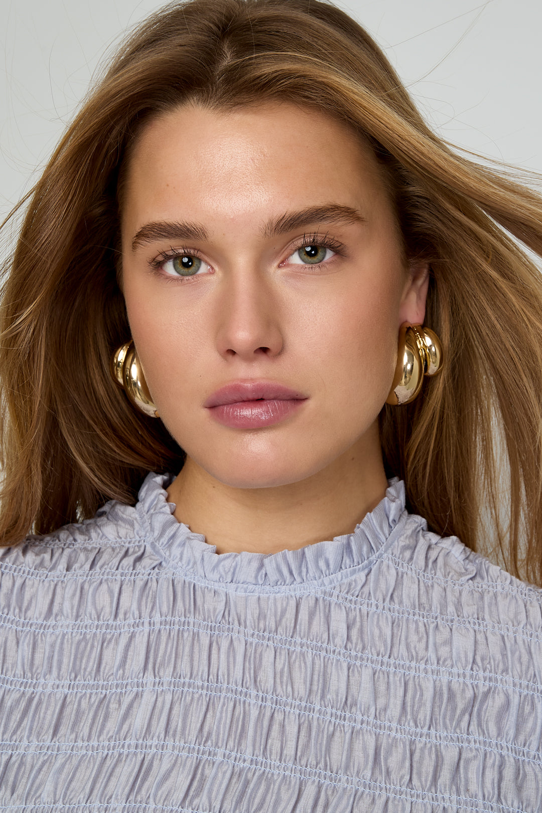 Close-up front view of a woman wearing chunky gold hoop earrings with minimal makeup and light blue gathered blouse