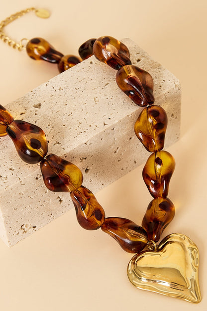 Close-up detail of amber acrylic beads and gold heart pendant resting on beige textured surface