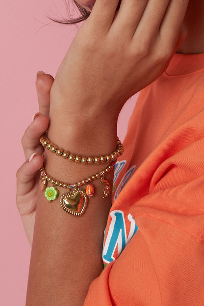 Model wearing gold charm bracelet and gold beaded bracelet on wrist with orange top and pink background