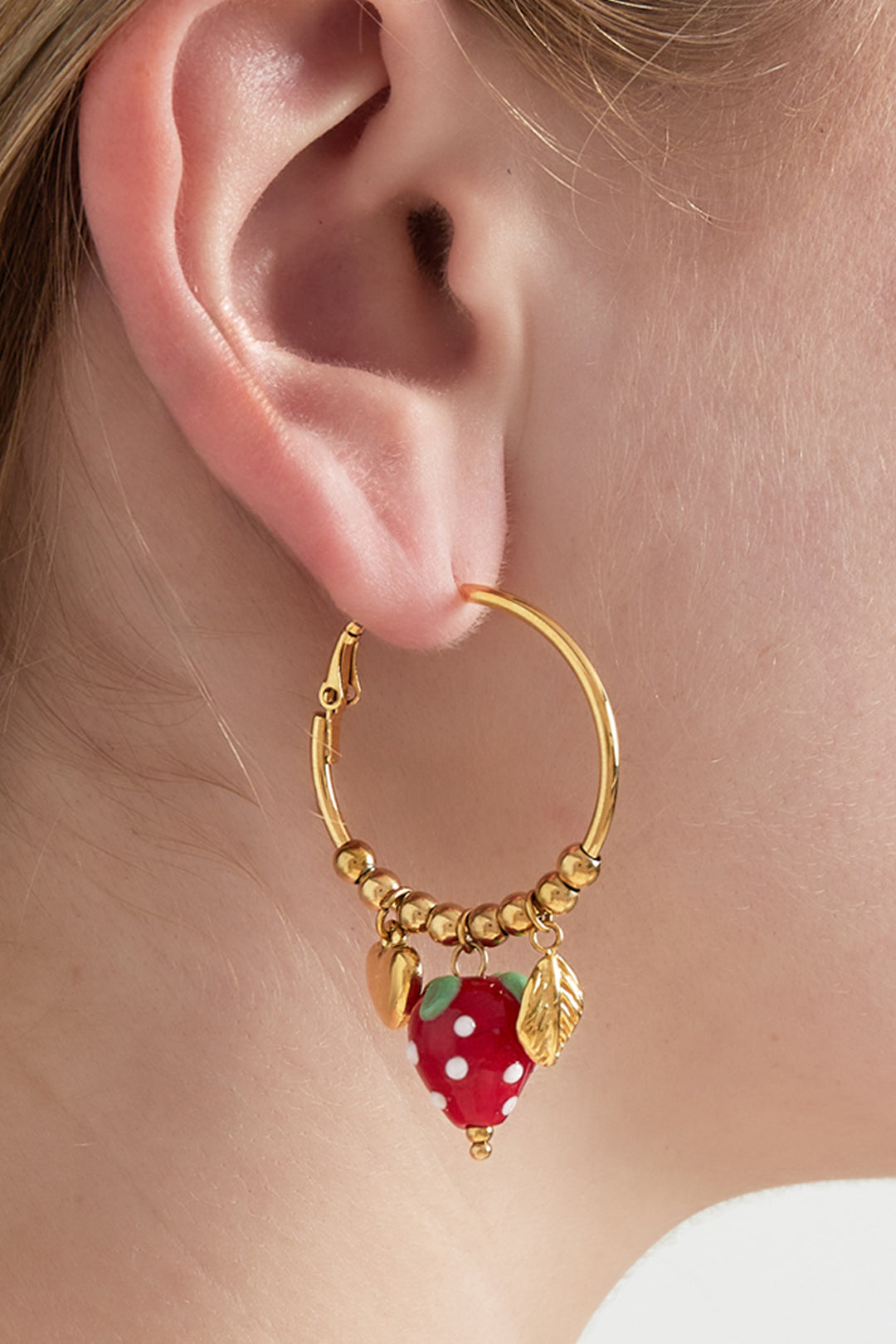 Close-up of a woman wearing gold-plated hoop earring with red enamel strawberry and gold leaf charms