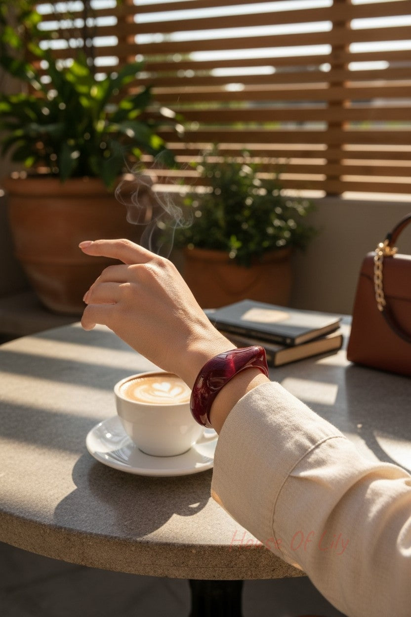 A smooth, translucent deep red acrylic statement bangle worn on a wrist at a cafe table with a steaming coffee.