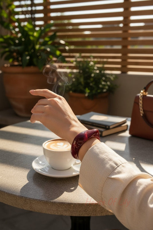 A smooth, translucent deep red acrylic statement bangle worn on a wrist at a cafe table with a steaming coffee.