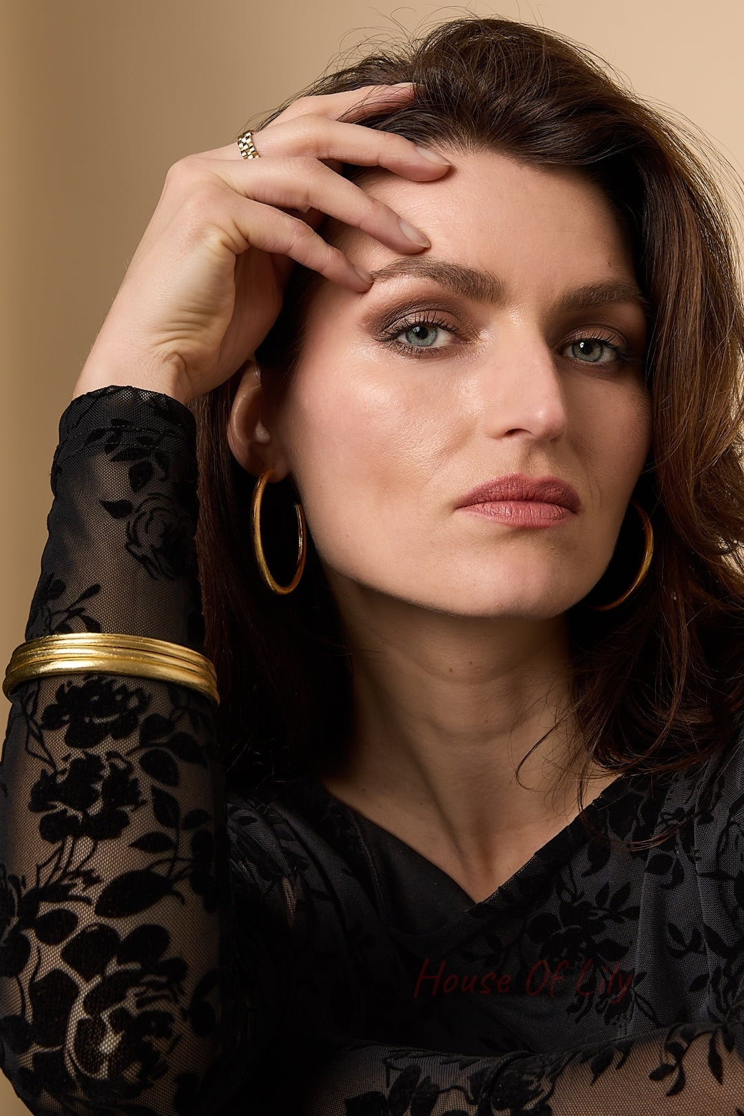 Woman wearing matching gold circle hoop earrings and several gold bangles stacked, dressed in sheer black floral top, posing with hand near face