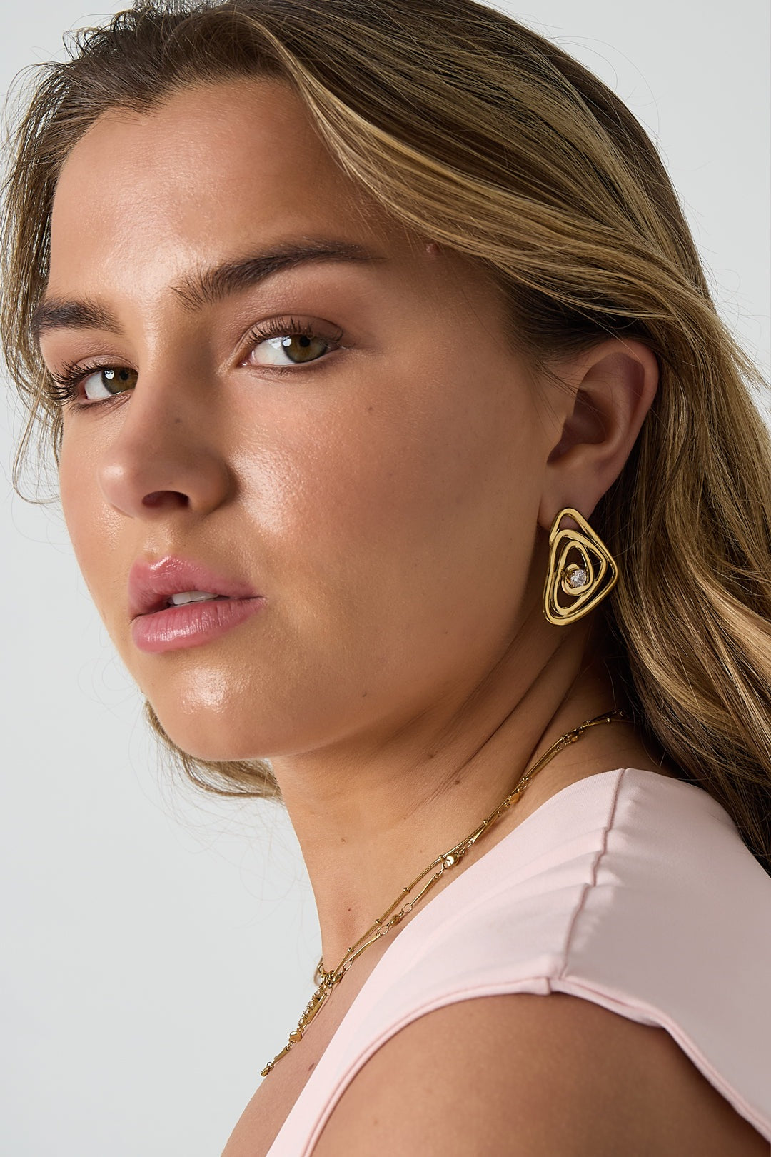 Close-up of a woman wearing gold spiral earrings with rhinestone, styled with delicate gold necklace and light pink top