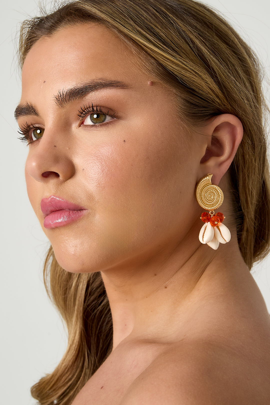 Portrait side angle of woman with gold spiral shell earrings featuring orange glass beads and white cowry shells