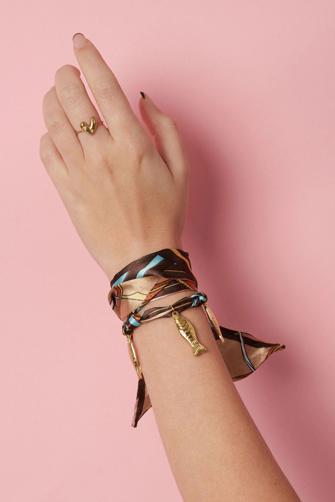 Close-up of a woman's wrist wearing the brown and turquoise silk wrap bracelet with gold fish charms against pink background