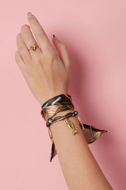 Close-up of a woman's wrist wearing the brown and turquoise silk wrap bracelet with gold fish charms against pink background