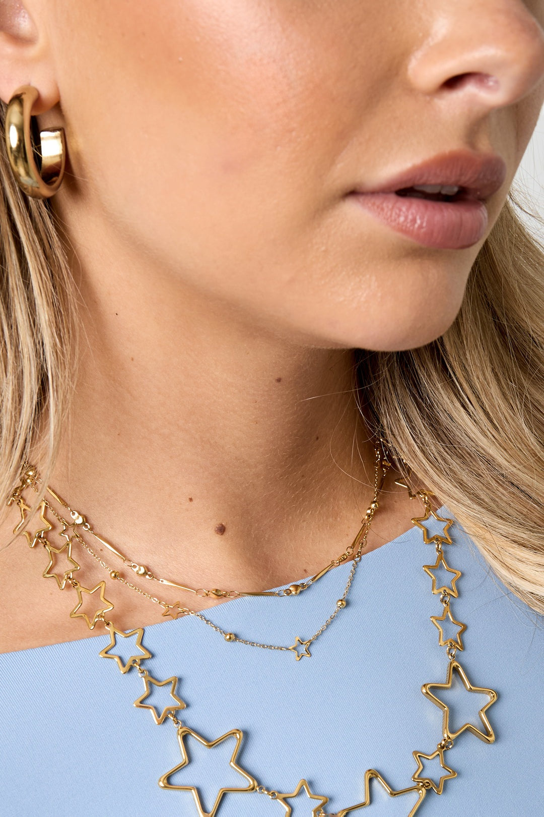Close-up of woman wearing layered gold star charm necklaces and gold hoop earrings with light blue top