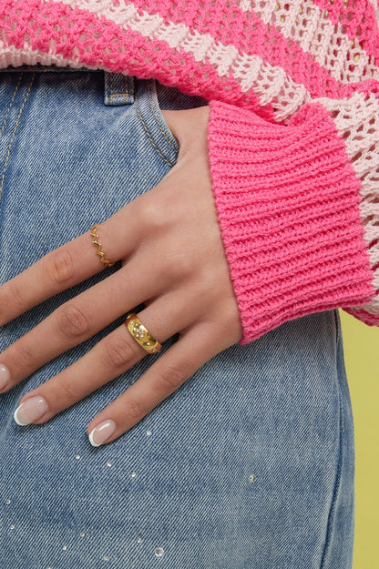 Woman's hand wearing the gold crystal accent ring on the middle finger and a wavy gold ring on the index finger, styled with pink knitwear and denim jeans with rhinestone details