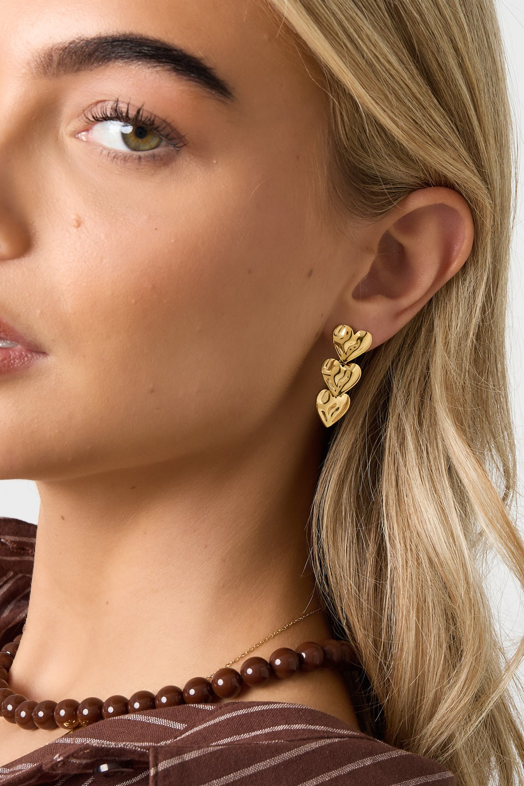 Close-up of a woman wearing gold triple heart dangle earrings styled with a brown beaded necklace
