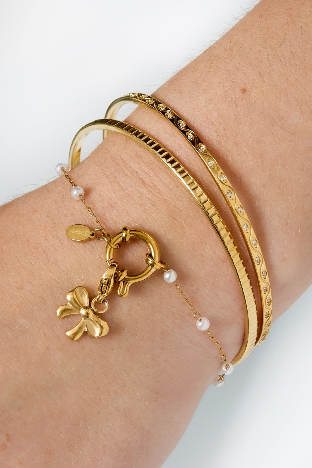 Close-up of wrist wearing three gold bracelets including delicate pearl chain bracelet with toggle clasp and bow charm