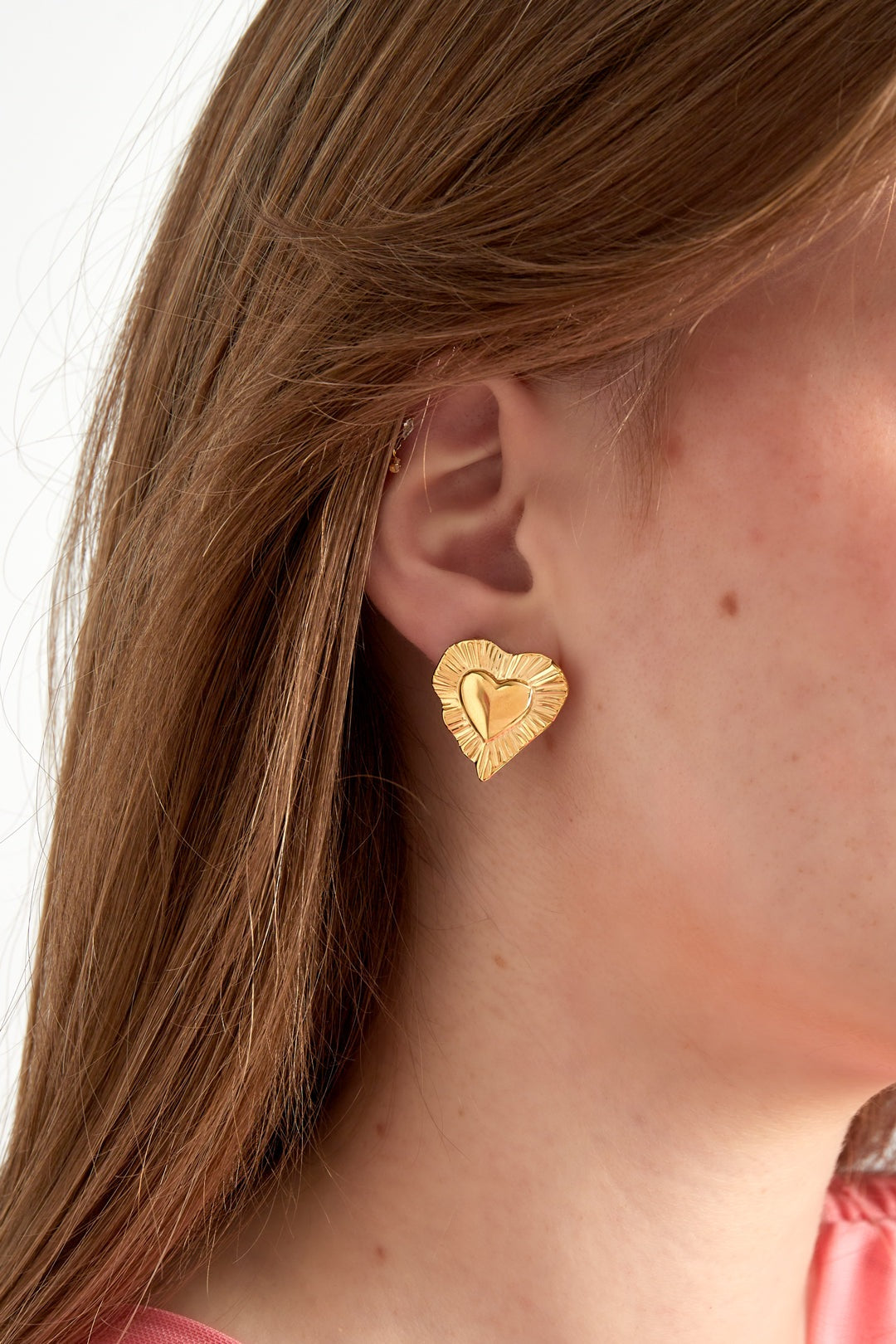 Close-up of gold radiant heart stud earring worn by woman with light brown hair and pink top