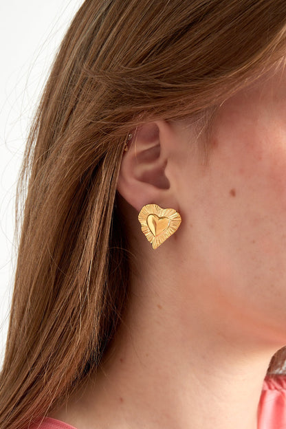 Close-up of gold radiant heart stud earring worn by woman with light brown hair and pink top