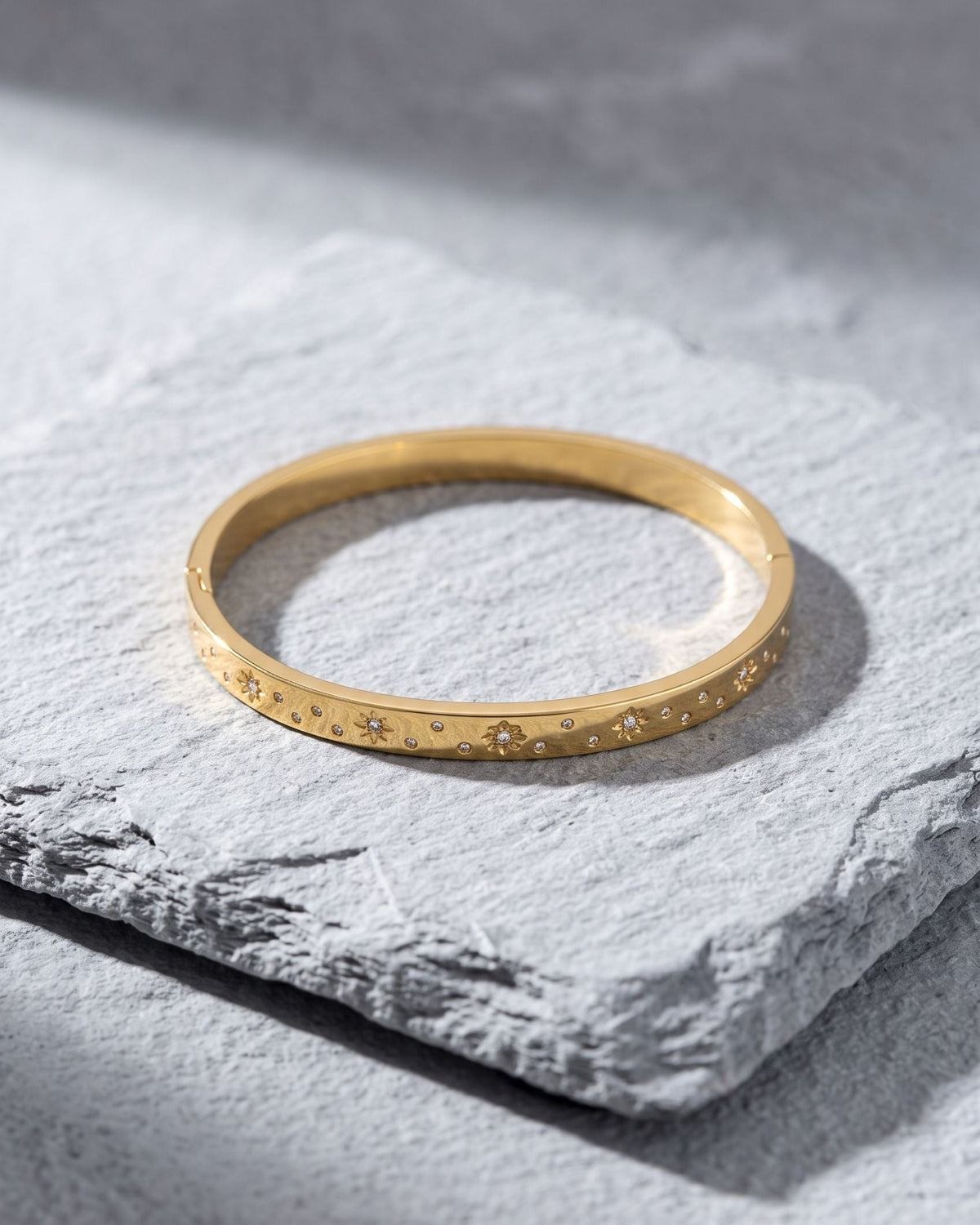 Elegant gold-toned stainless steel flower bangle resting on a textured light grey slate surface.
