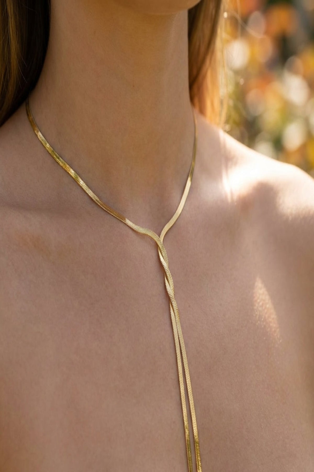 Close-up of a delicate gold twist necklace with tassel strands on a model's neck, highlighting polished finish and minimalist design in natural sunlight.
