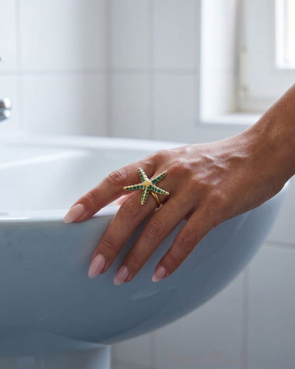 Elegant adjustable starfish ring with green enamel on a model's finger resting on a white porcelain sink edge.