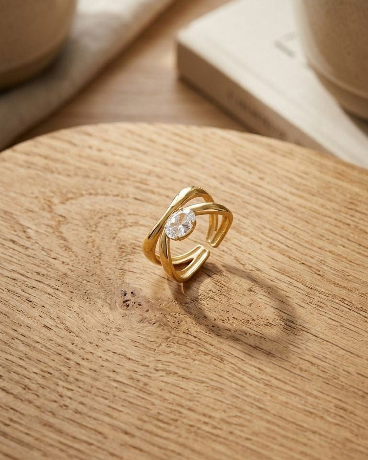 Minimal scene of Twisted Oval Crystal ring with gold-tone steel on textured wood, captured from above with warm lighting.