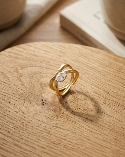 Minimal scene of Twisted Oval Crystal ring with gold-tone steel on textured wood, captured from above with warm lighting.