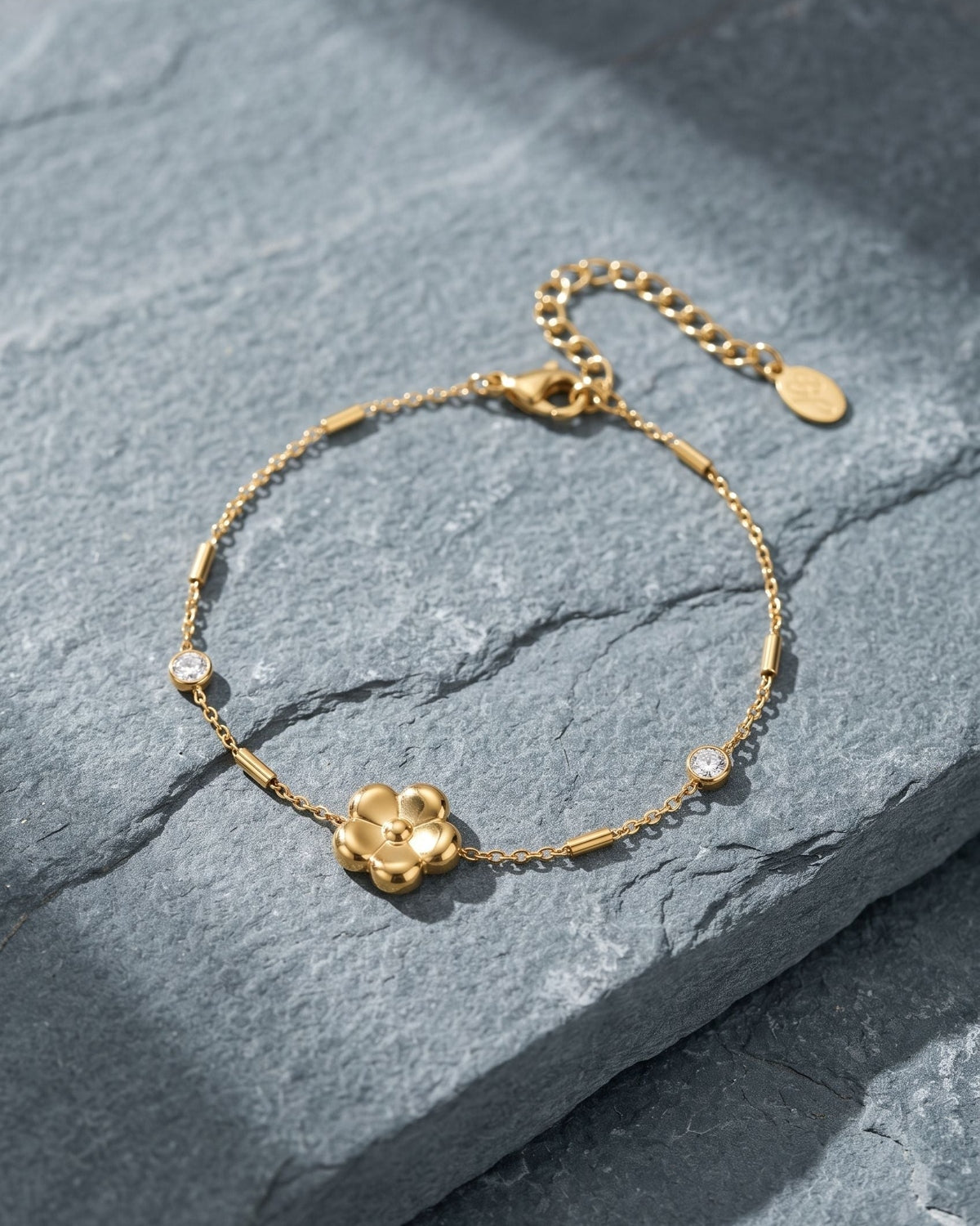 Delicate stainless steel flower charm bracelet with sparkling zircon stones resting on a textured grey stone surface.