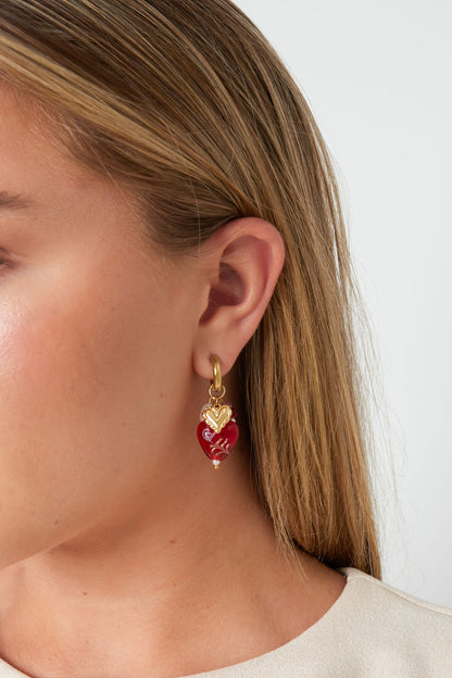 Woman wearing a gold hoop earring featuring a red heart charm with floral design and a gold mini heart pendant