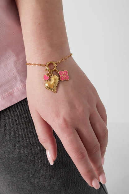 Woman’s wrist wearing gold-plated charm bracelet with heart, pink enamel heart, and pink flower charms, styled with pink top and dark pants