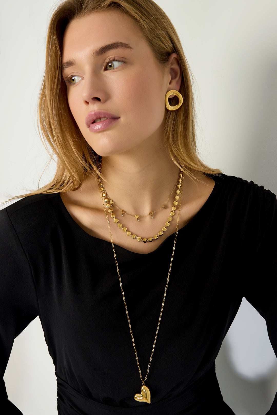 Model wearing layered gold necklaces including heart link chain, a delicate heart charm necklace, and long pendant necklace with gold textured earrings