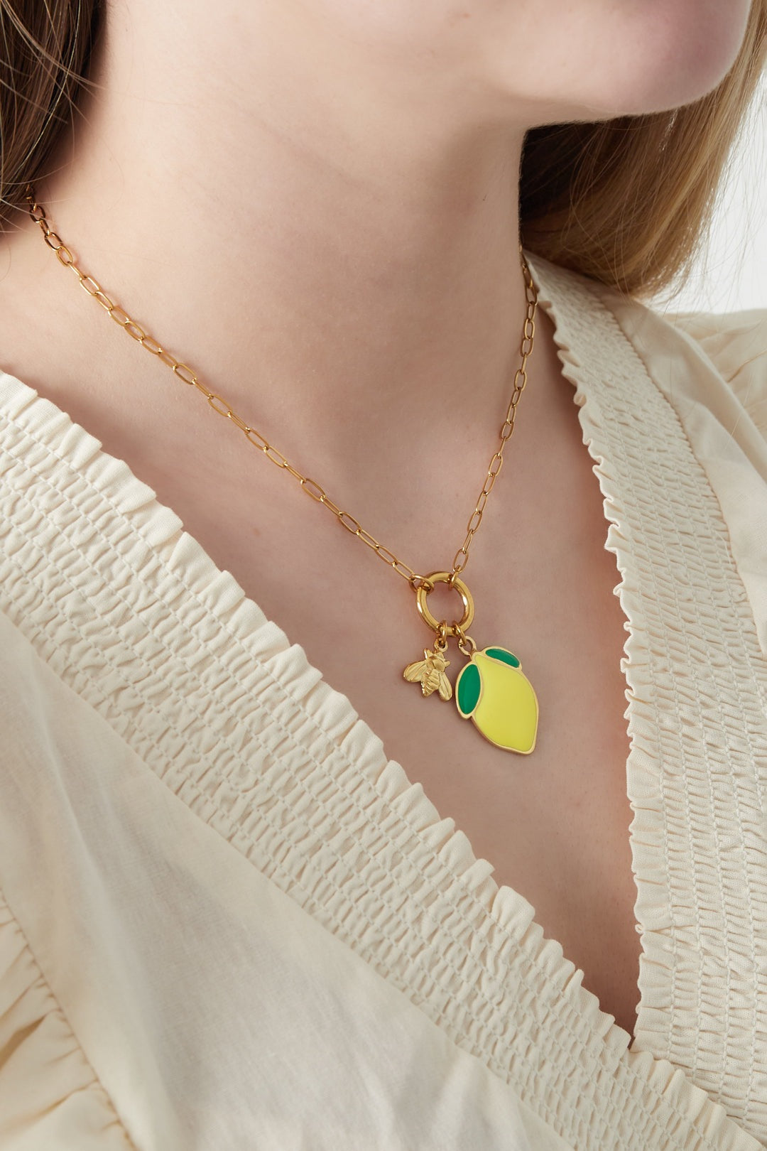 Close-up of a woman wearing a gold chain necklace with a yellow lemon charm and bee detail, paired with a cream blouse