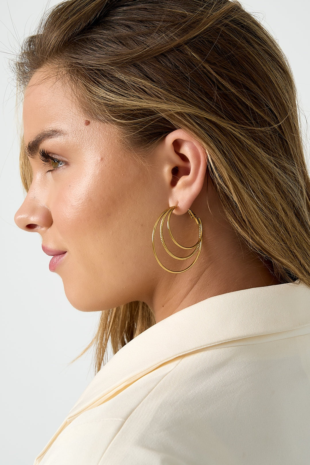Side profile of woman wearing gold triple layered hoop earrings showcasing elegant minimalistic layered design against ivory blazer
