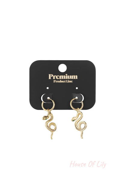 Gold hoop earrings featuring dangling snake charms with crystal-accented bodies and green stone eyes on a black card.