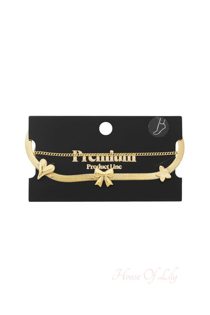 Dainty gold double-chain anklet featuring a flat snake chain with heart, bow, and flower charms on a display card.