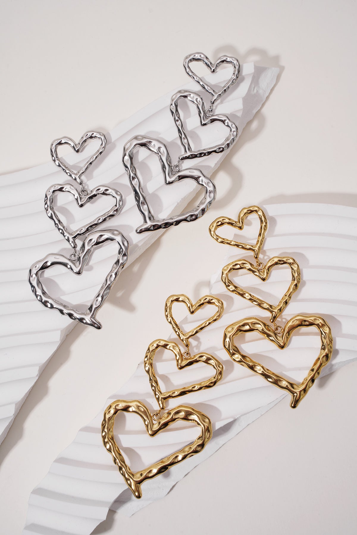 Triple Heart Energy earrings in gold and silver, featuring three graduated, hammered-metal heart shapes in a drop style.