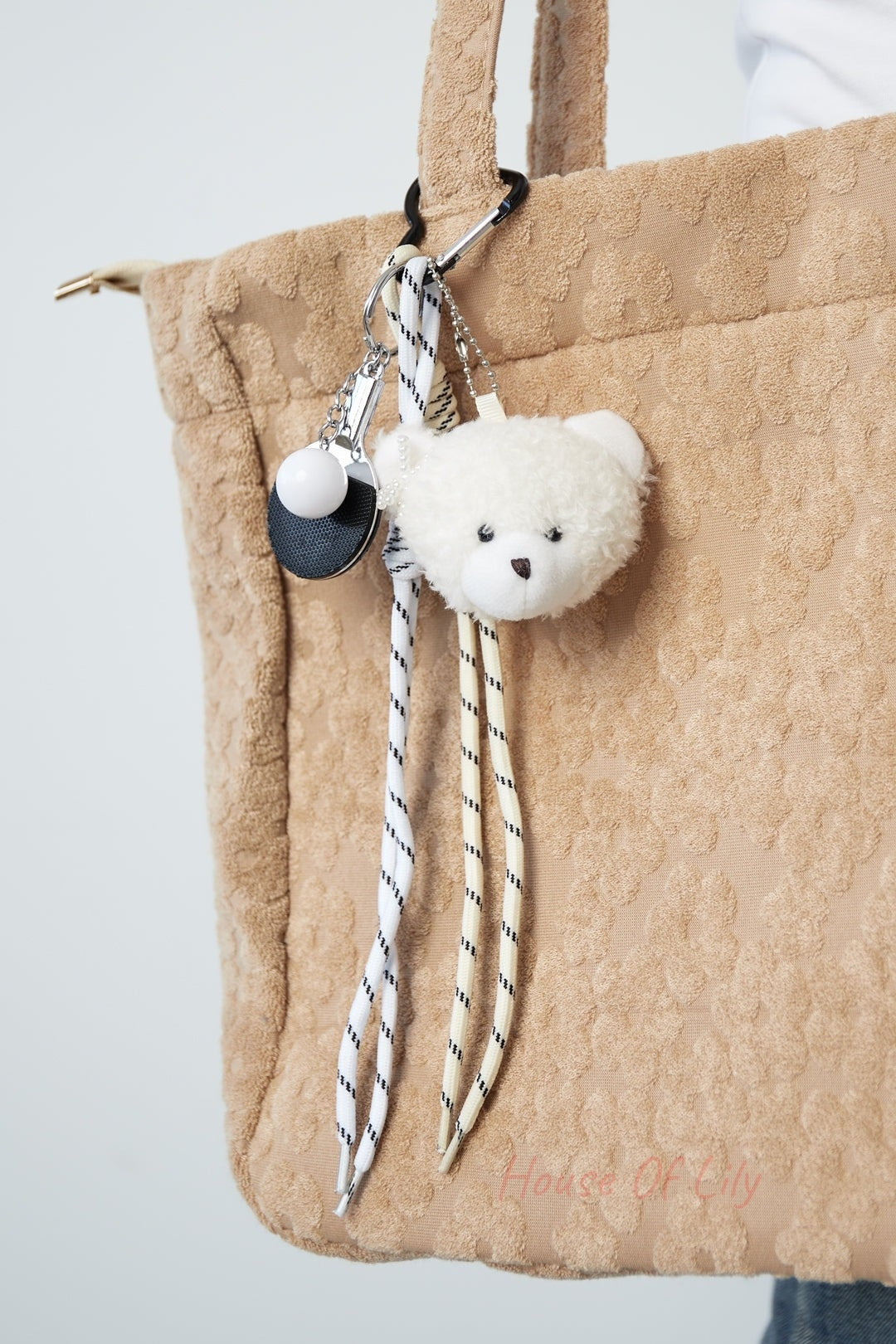 Close-up of plush bear bag charm clipped on a beige textured tote bag, showing the soft bear head, striped laces, mini ping pong paddle, and heart-shaped clip.