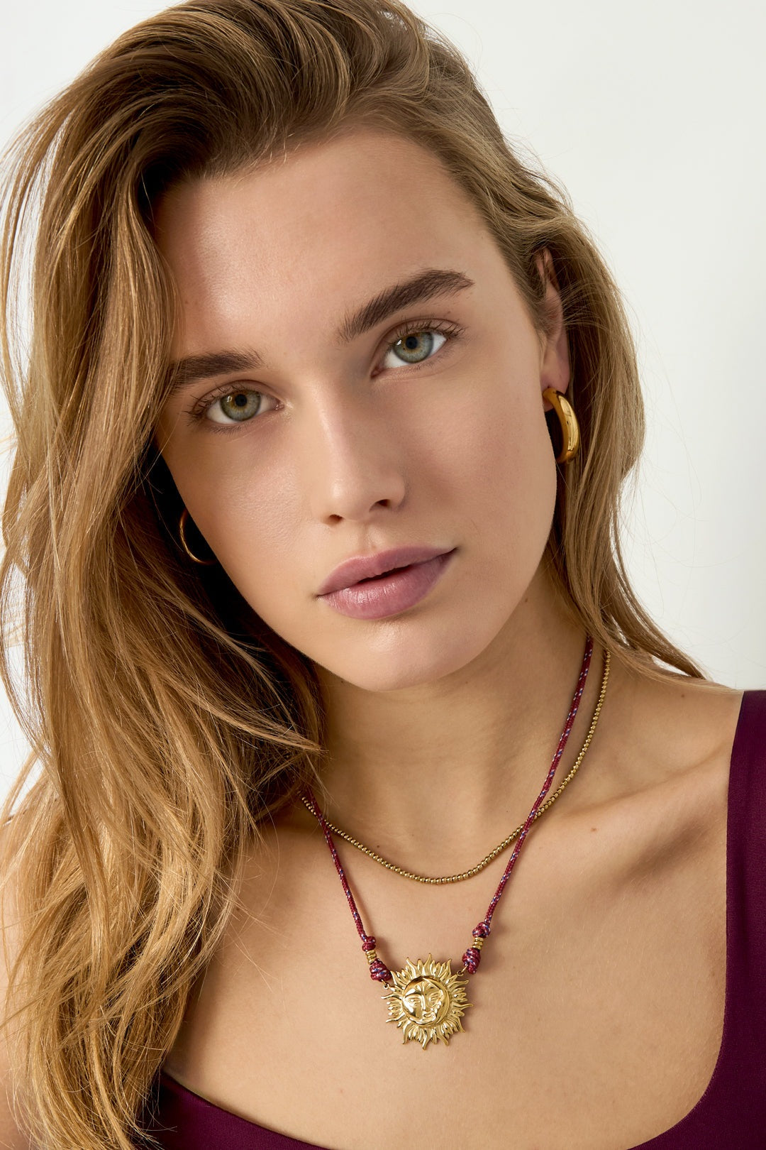 Model wearing gold-tone sun pendant necklace on red rope cord with a simple purple top and gold hoop earrings