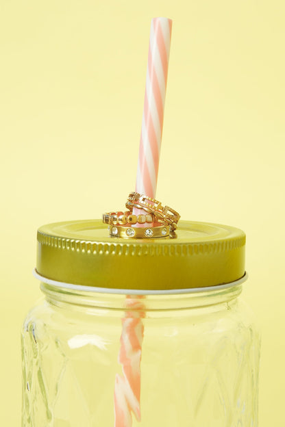 Three gold rings with beaded and gemstone designs stacked on a pink-striped straw in a green lid mason jar on yellow background
