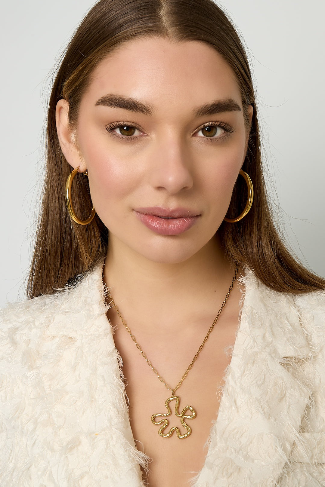 Close-up of a woman wearing the gold floral pendant necklace paired with gold hoop earrings and a textured white jacket