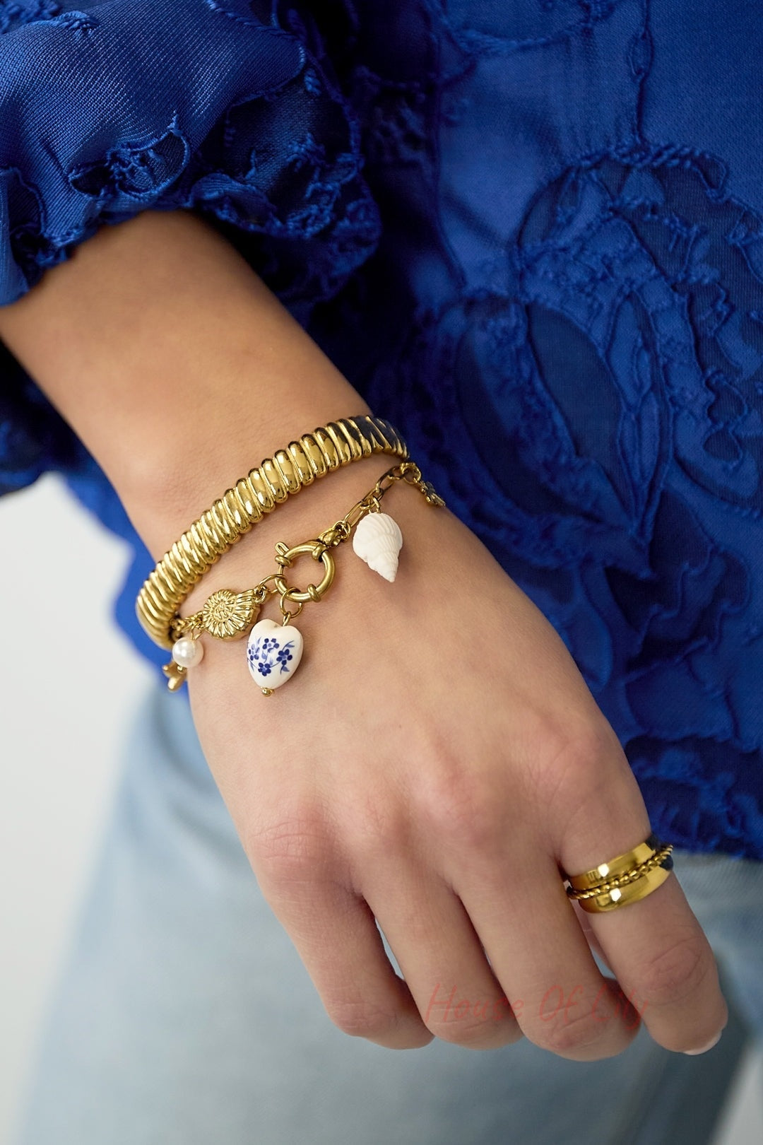 Woman's wrist wearing gold ribbed bangle with charm bracelet and rings wearing blue lace top and jeans