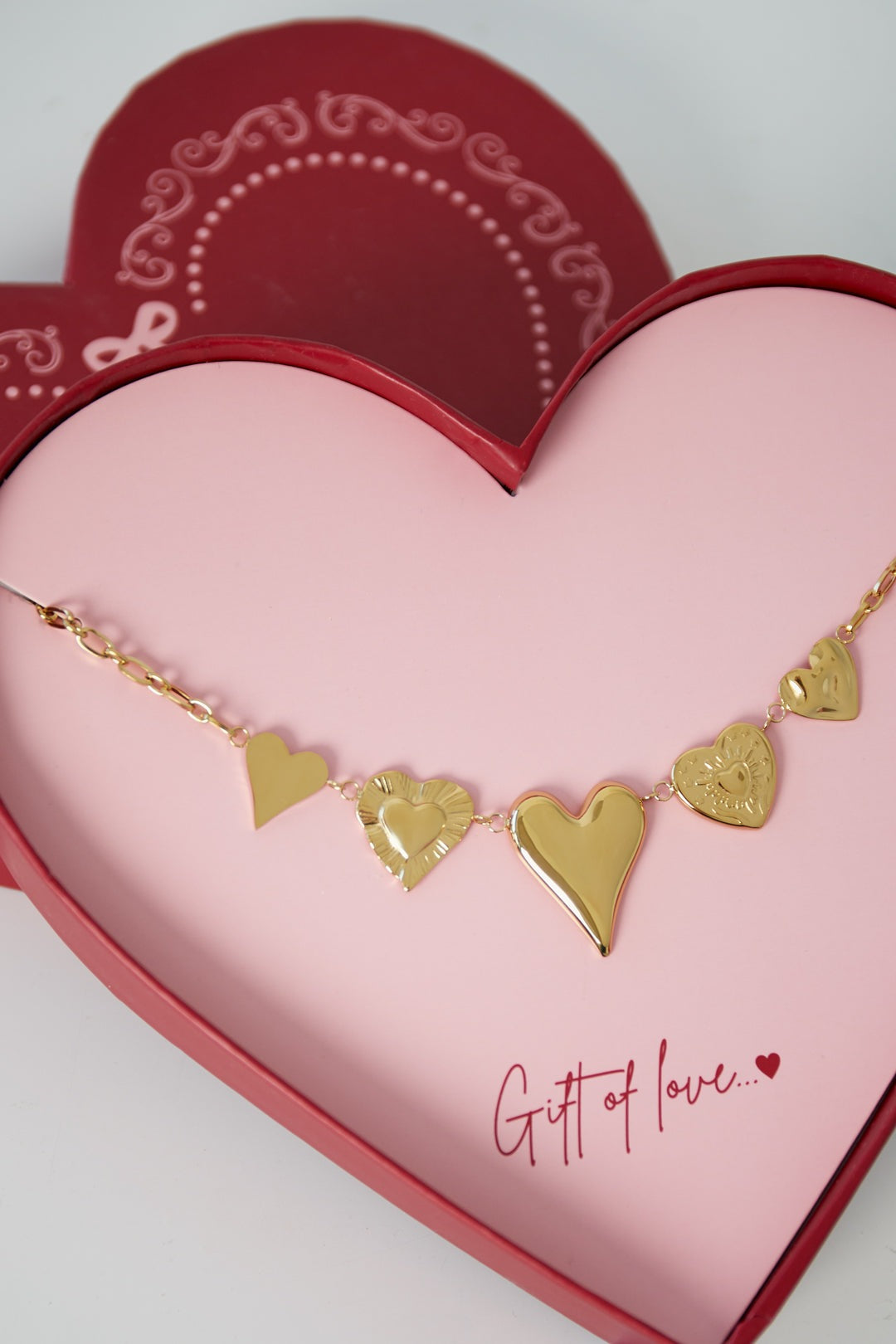 Close-up view of the gold heart charm necklace showing detailed textures on each heart pendant inside a pink and red heart-shaped box.