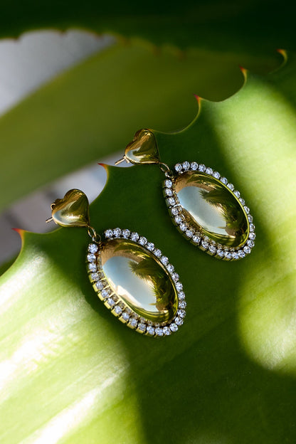 Main Character Drip earrings: gold, oval-shaped drops with crystal halos and heart studs, resting on a green leaf.