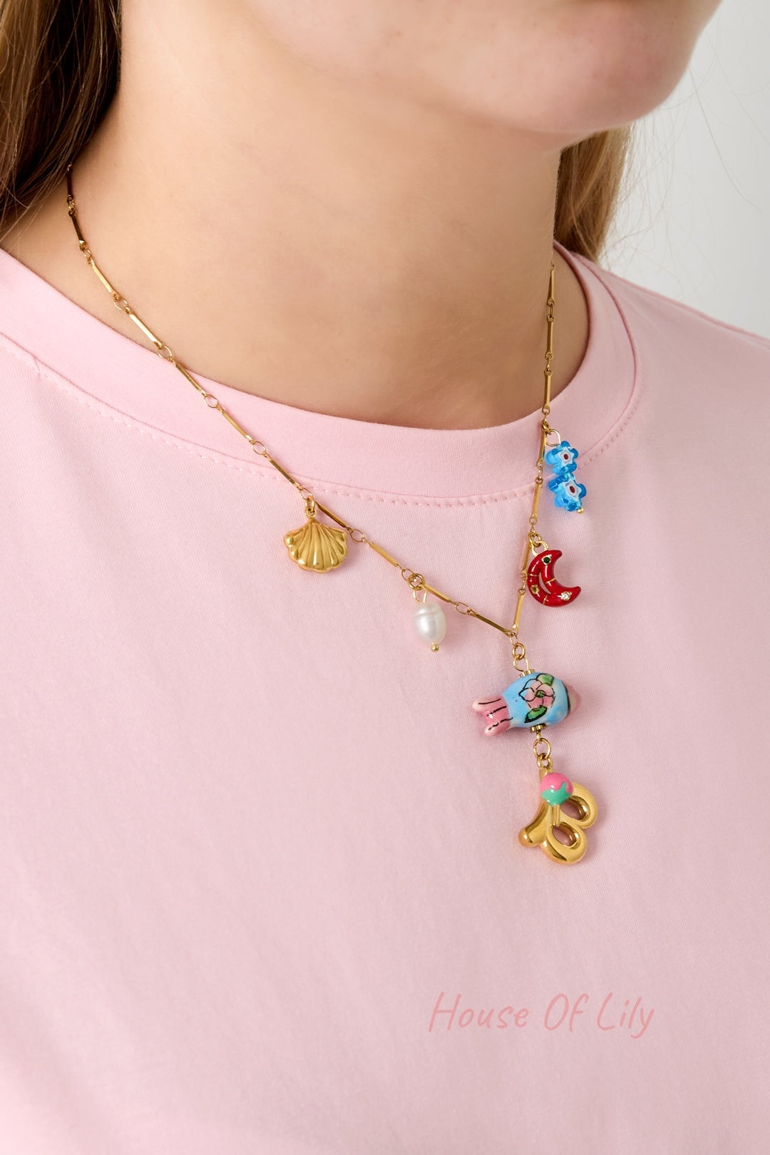 Model wearing a gold charm necklace with gold seashell, white pearl, red crescent moon, blue flowers, and gold letter B with pink flower pendant