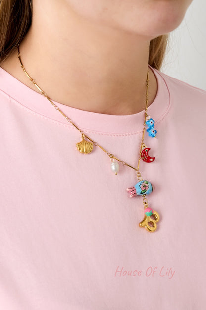 Model wearing a gold charm necklace with gold seashell, white pearl, red crescent moon, blue flowers, and gold letter B with pink flower pendant