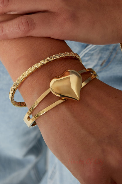 Close-up of wrist wearing a shiny gold double-band heart bangle with a second textured gold bracelet, styled with casual denim