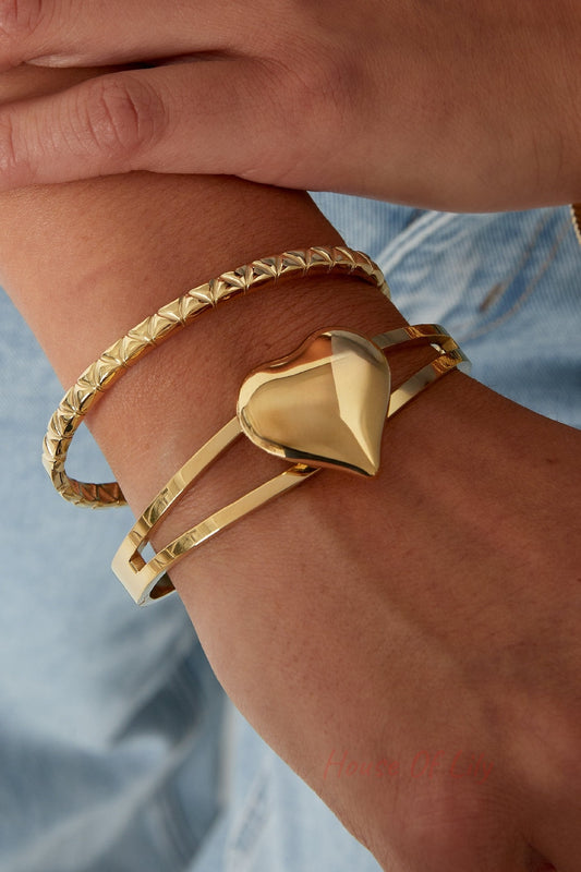 Close-up of wrist wearing a shiny gold double-band heart bangle with a second textured gold bracelet, styled with casual denim