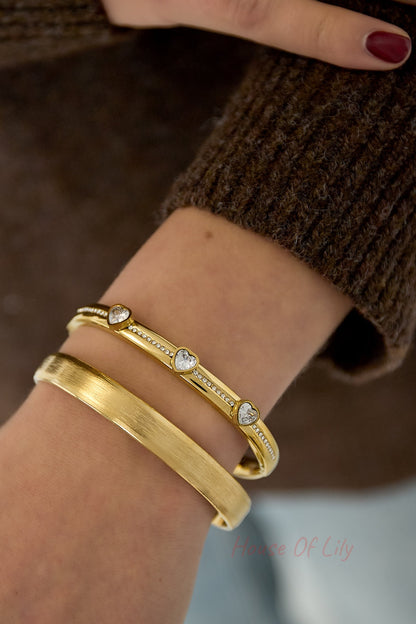 Woman's wrist wearing gold tone heart crystal bangle with a second matte gold bracelet alongside brown knit sweater