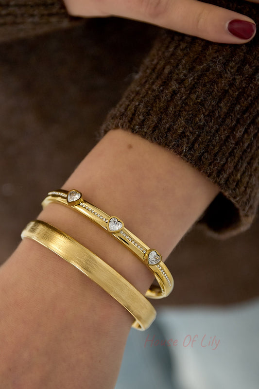 Woman's wrist wearing gold tone heart crystal bangle with a second matte gold bracelet alongside brown knit sweater